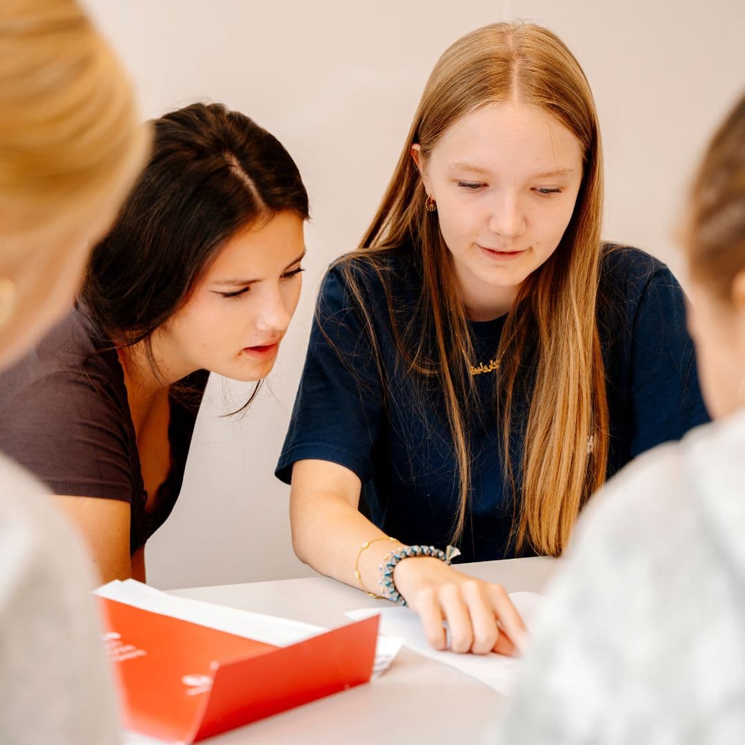 To piger forbereder sig til deres 9. klasseprøve i dansk og læser papirer sammen ved et bord. Den ene peger på et ark, mens den anden læner sig frem, og der er yderligere to personer, der er slørede i forgrunden.
