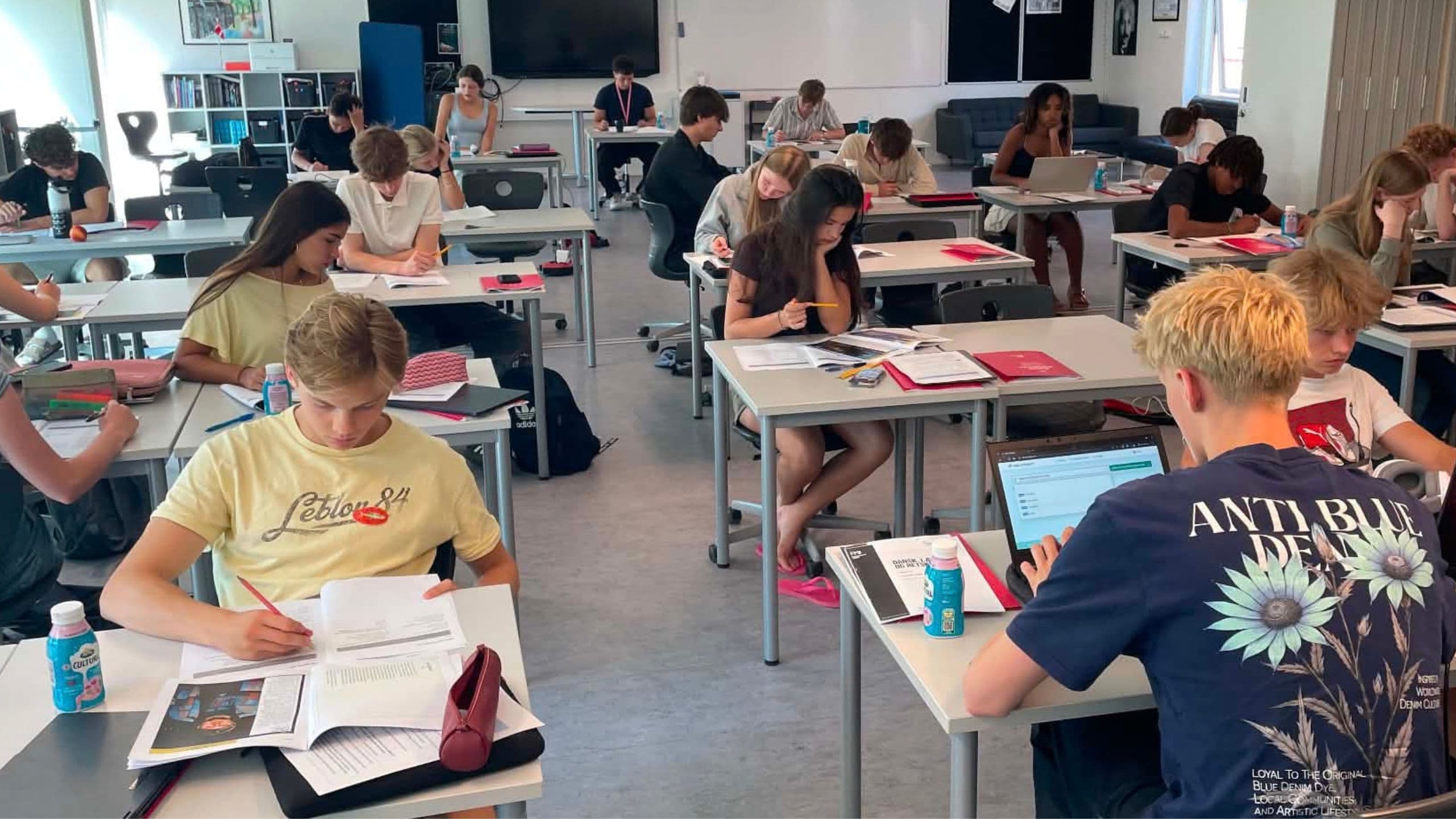 Eleverne sidder ved skrivebordene i et klasseværelse og arbejder på opgaver eller bruger bærbare computere. Klasseværelset er lyst, og der står stole, borde og skoleartikler spredt rundt omkring. De fleste elever ser ud til at være koncentrerede om deres arbejde.