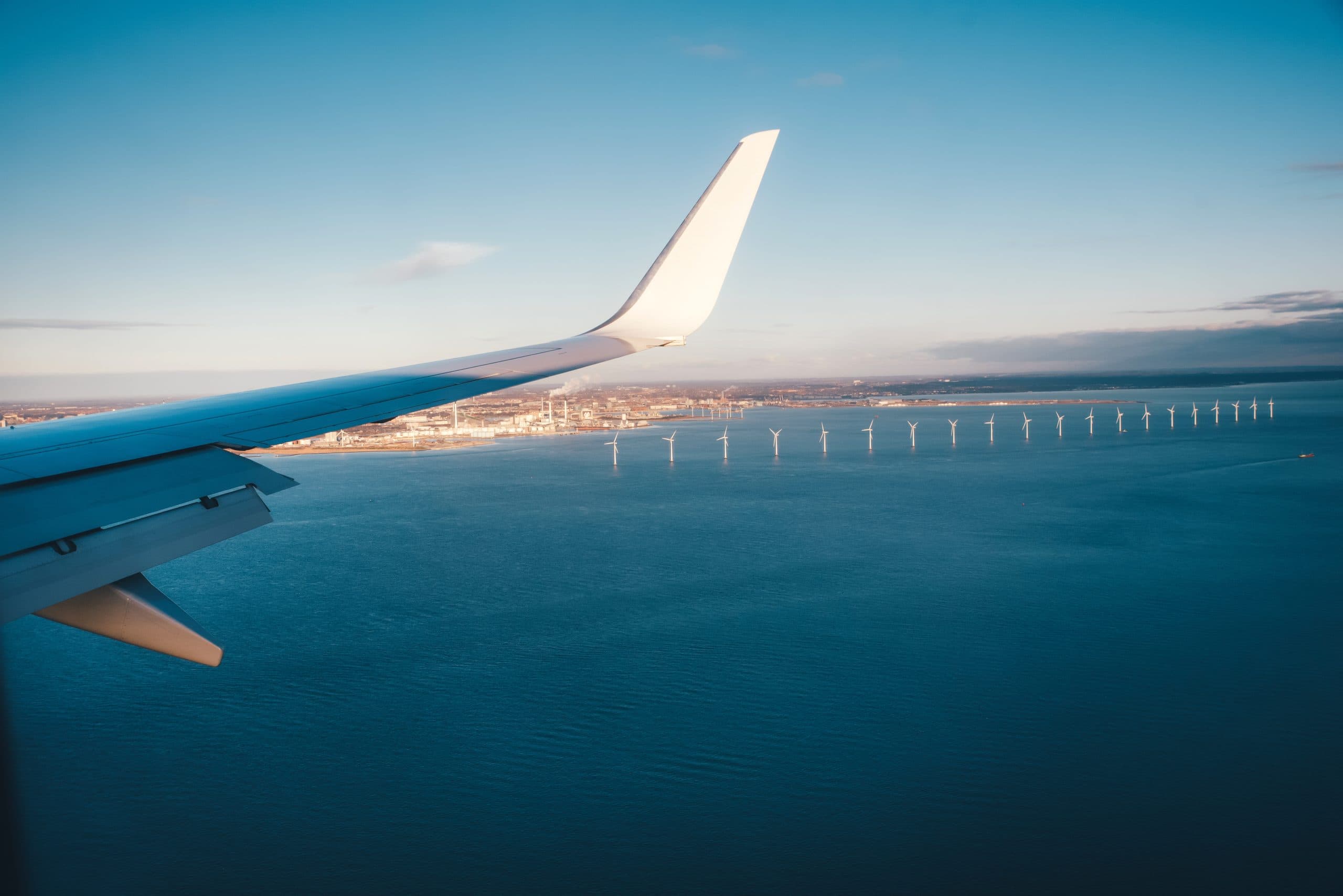 Udsigt fra et flyvindue, der viser vingen over et stort vandområde med vindmøller i det fjerne og en by langs kysten under en klar blå himmel.