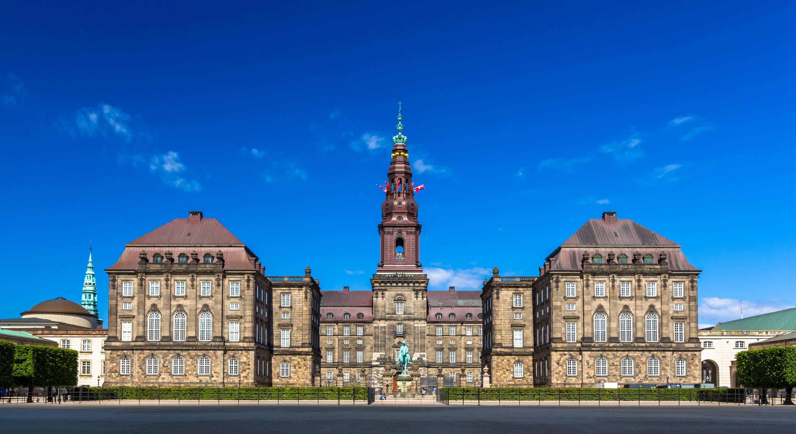 En storslået historisk bygning med et centralt tårn og to symmetriske fløje mod en lyseblå himmel; en statue står foran, og klippede hække afgrænser forgrunden.