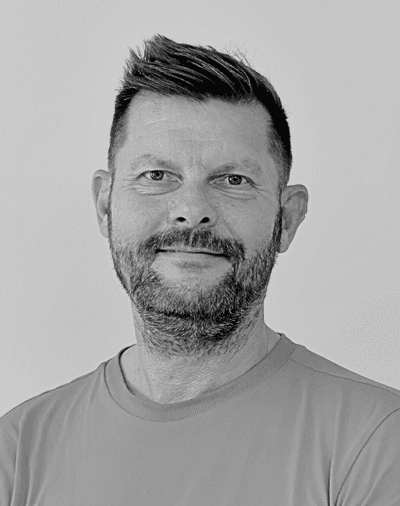 Sort-hvidt portræt af en mand med kort, stylet hår og skæg, iført en ensfarvet T-shirt, der smiler let, mens han ser direkte ind i kameraet foran en lys baggrund.