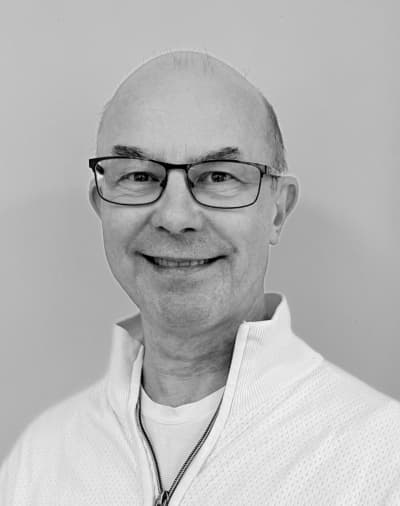En smilende ældre mand med briller og høj pande, iført en lys trøje med lynlås, der står foran en ensfarvet baggrund. Billedet er i sort-hvid.