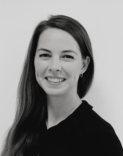 Et sort-hvidt portræt af en kvinde med langt, glat hår, der smiler, iført en mørk bluse og en lille ringørering, stående foran en ensfarvet lys baggrund.