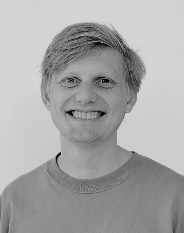 En person med lyst hår og et bredt smil har en ensfarvet T-shirt med rund hals på. Billedet er i sort-hvid, og baggrunden er ensfarvet og lys.