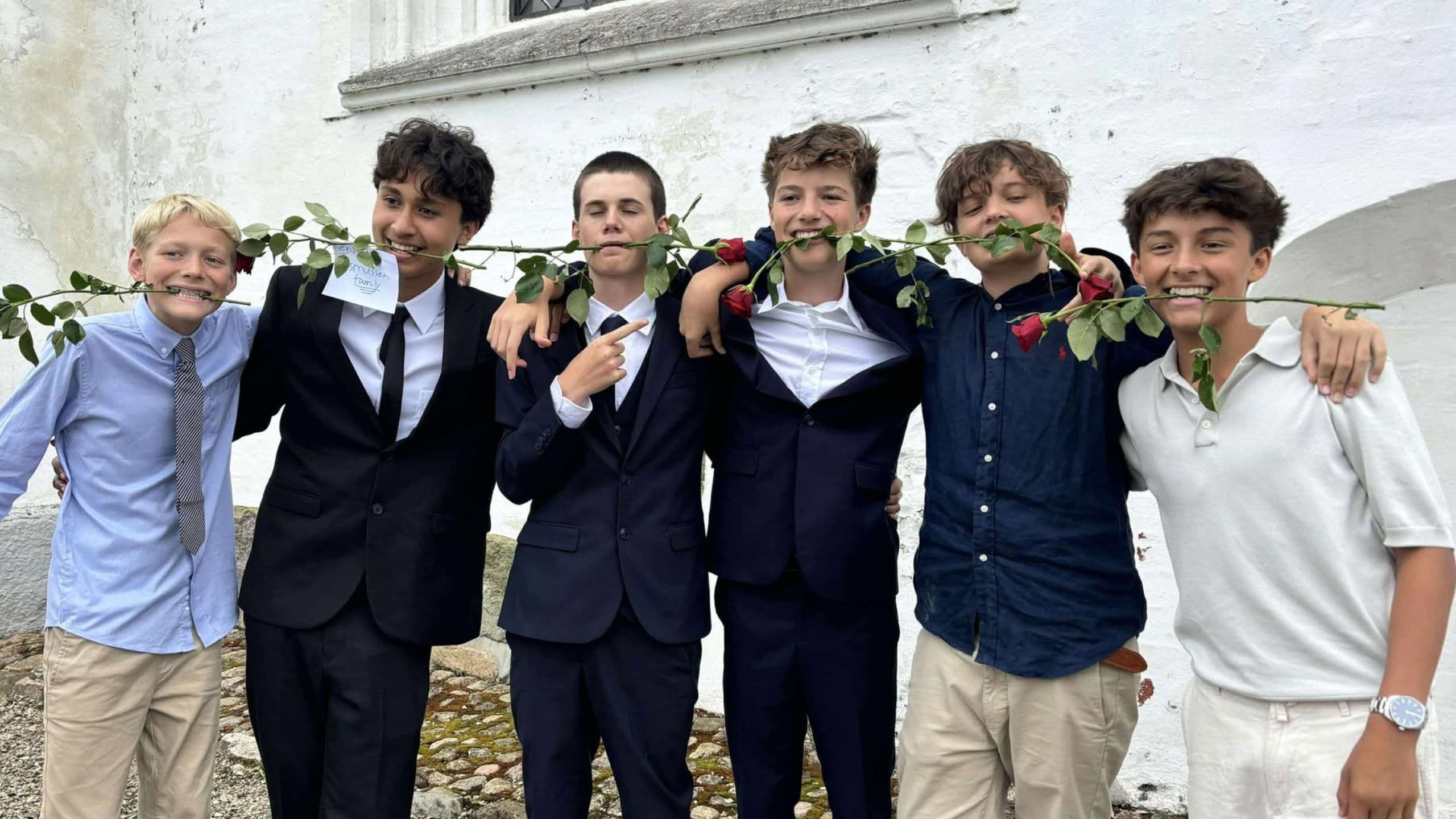 Seks drenge står og smiler foran en hvid væg; fire af dem er iført jakkesæt og har roser i munden, mens to har afslappede skjorter på. De poserer alle legesygt med armene om hinanden.