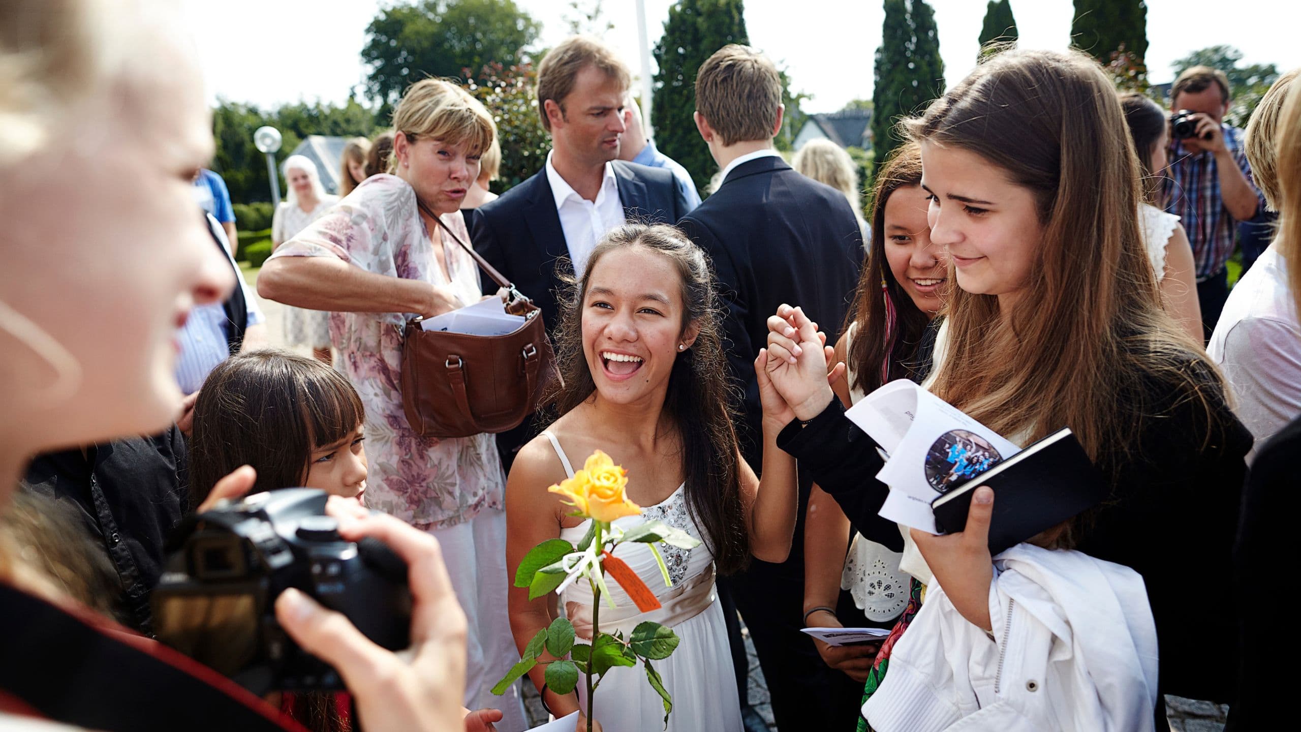 En gruppe unge står udenfor, smiler og fejrer. En pige i midten holder en gul rose og ser glad ud. Folk omkring hende snakker, tager billeder og holder aviser eller programmer i hånden. Der er træer i baggrunden.