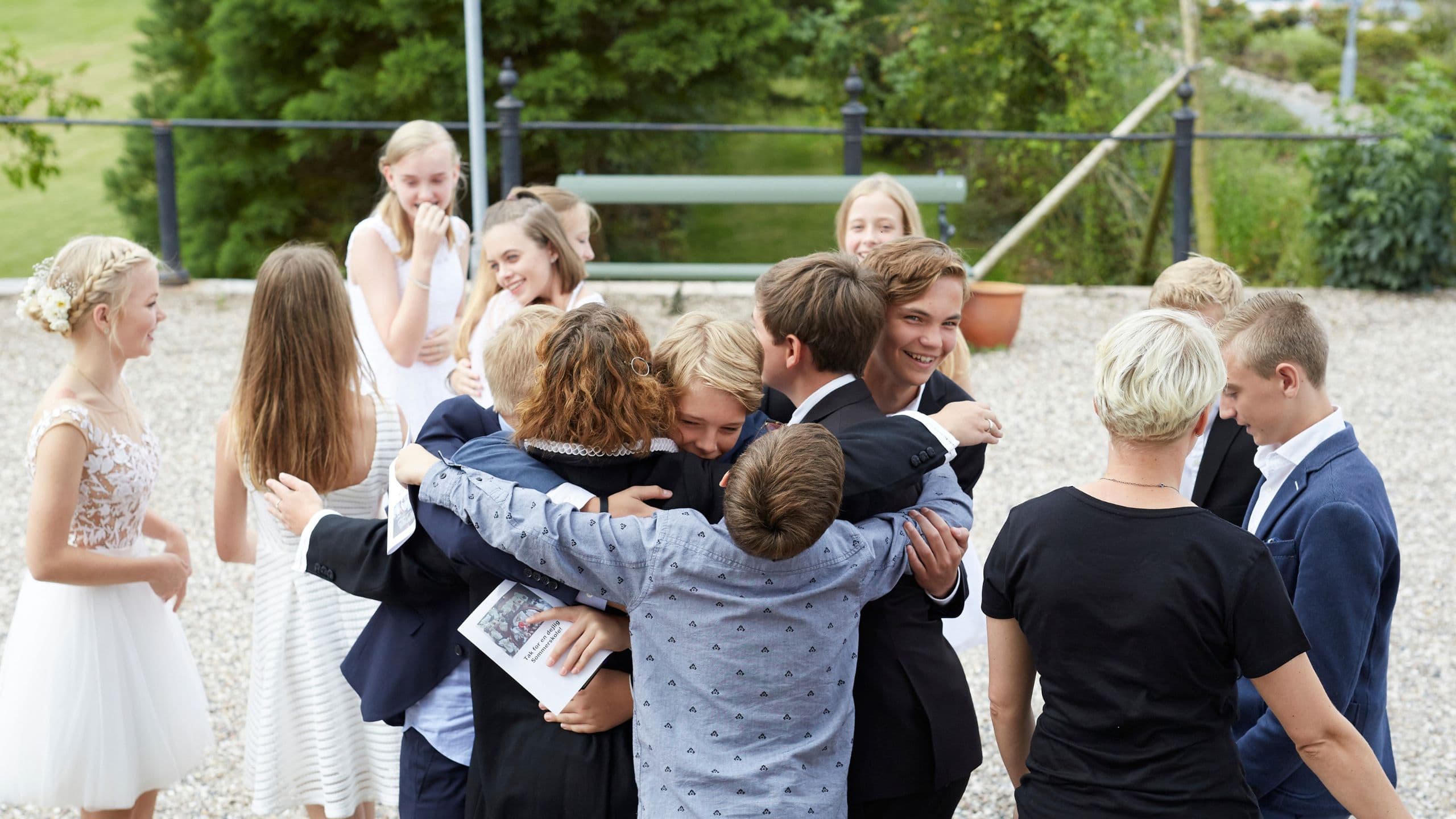 En gruppe teenagere, hvoraf nogle er klædt i jakkesæt og kjoler, omfavner hinanden og smiler udendørs i en festlig atmosfære ved en dansk konfirmation i Danmark, mens andre står og snakker i baggrunden.