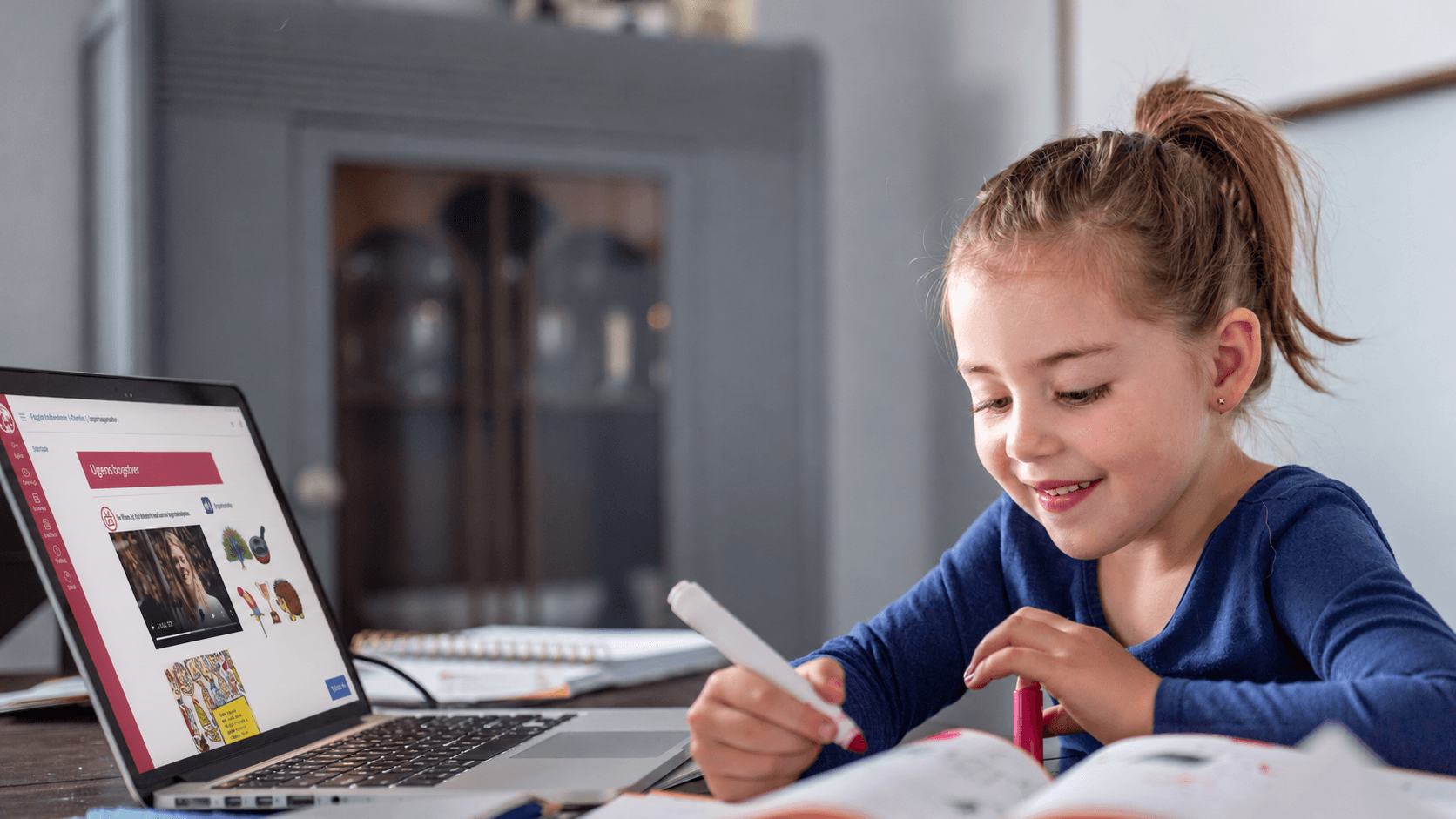 En ung pige smiler, mens hun bruger en tuschpen til at skrive i en online-arbejdsbog til dansk folkeskole ved siden af en åben bærbar computer.
