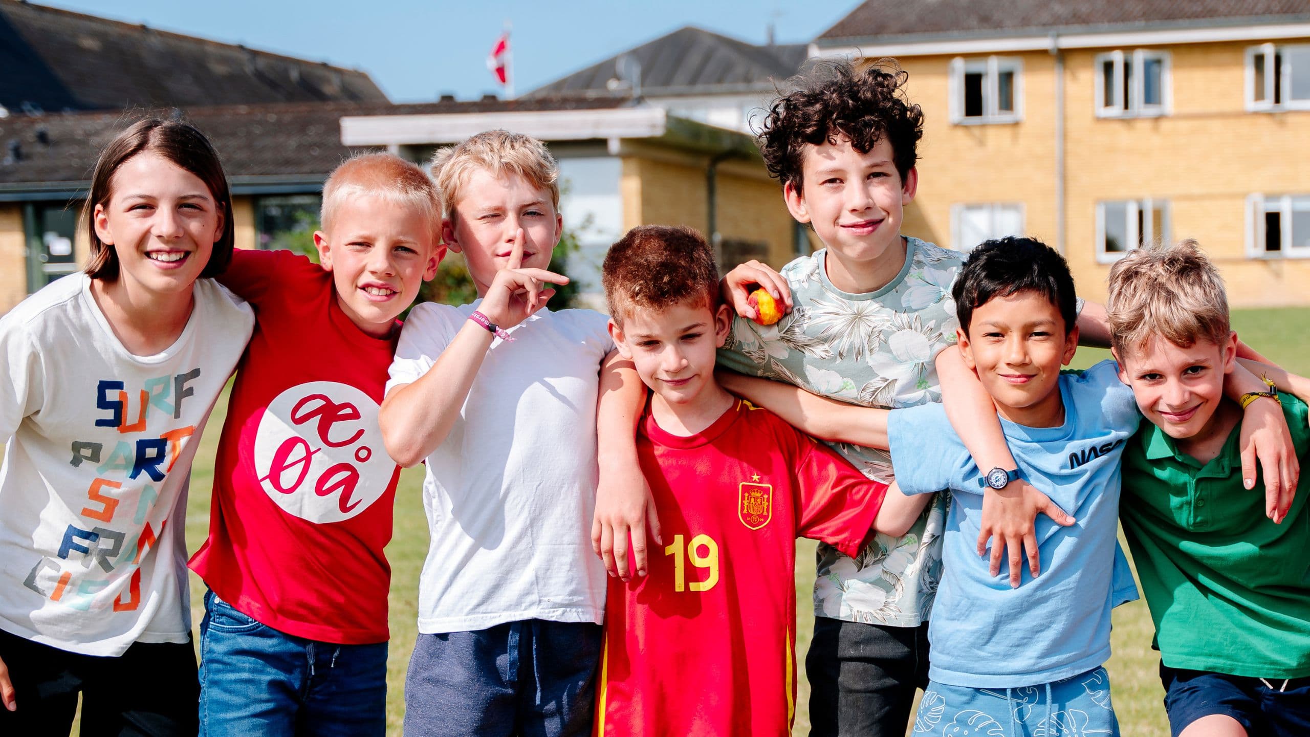Syv børn står på række udendørs med armene om hinandens skuldre og smiler til kameraet, mens de deltager i Den Danske Sommerskole. Få rabat på ophold som medlem af Danes Worldwide.