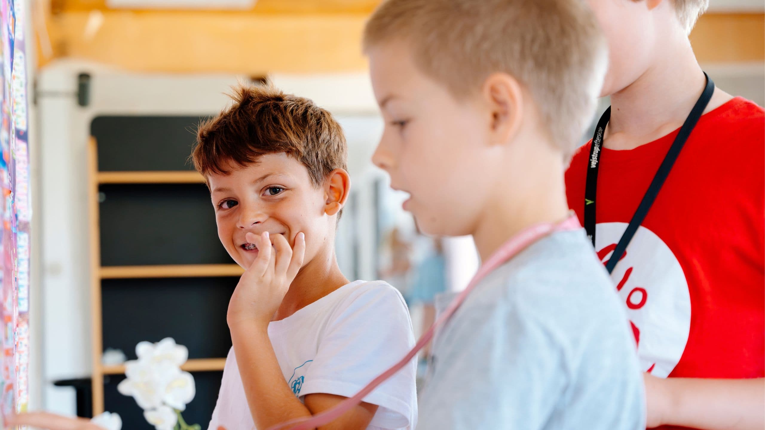Tre små drenge står sammen indenfor på en sommerskole for børn. En dreng i en hvid skjorte smiler til kameraet, mens de andre, klædt i gråt og rødt, kigger væk fra billedet. I baggrunden ses hylder og en hvid blomst.