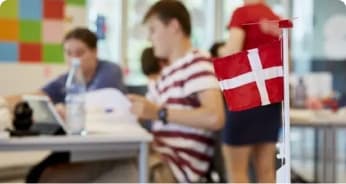 Et lille dansk flag på et skrivebord er i fokus, mens slørede elever arbejder i baggrunden i et farverigt, moderne klasseværelse.