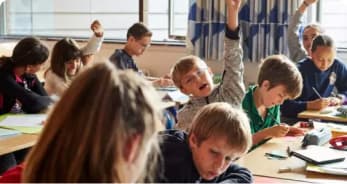 Et klasseværelse med unge elever, der sidder ved borde, nogle skriver eller læser, mens to drenge i midten ivrigt rækker hånden op for at svare på et spørgsmål.