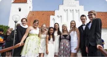 En gruppe formelt klædte unge mennesker står sammen og smiler foran en hvid kirke med rødt tegltag på en solrig dag.