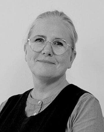 En kvinde med lyst hår, der er trukket tilbage, iført runde briller, en lys langærmet skjorte og en mørk ærmeløs vest, smiler blidt til kameraet mod en ensfarvet baggrund.