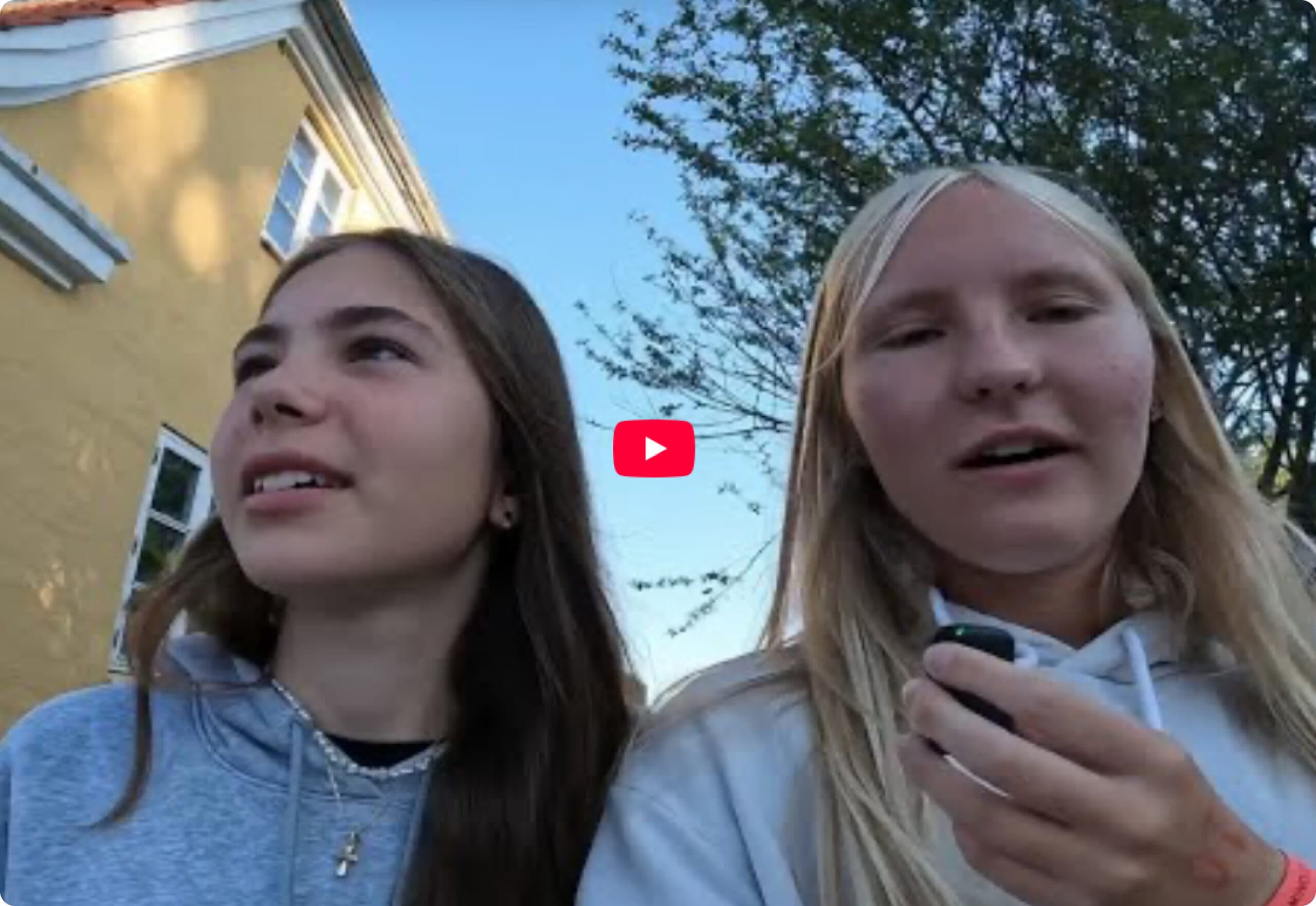 To teenagepiger med langt hår og hættetrøjer står udenfor i dagslys i nærheden af en gul bygning og træer. Den ene pige holder en lille genstand, og begge kigger i forskellige retninger.