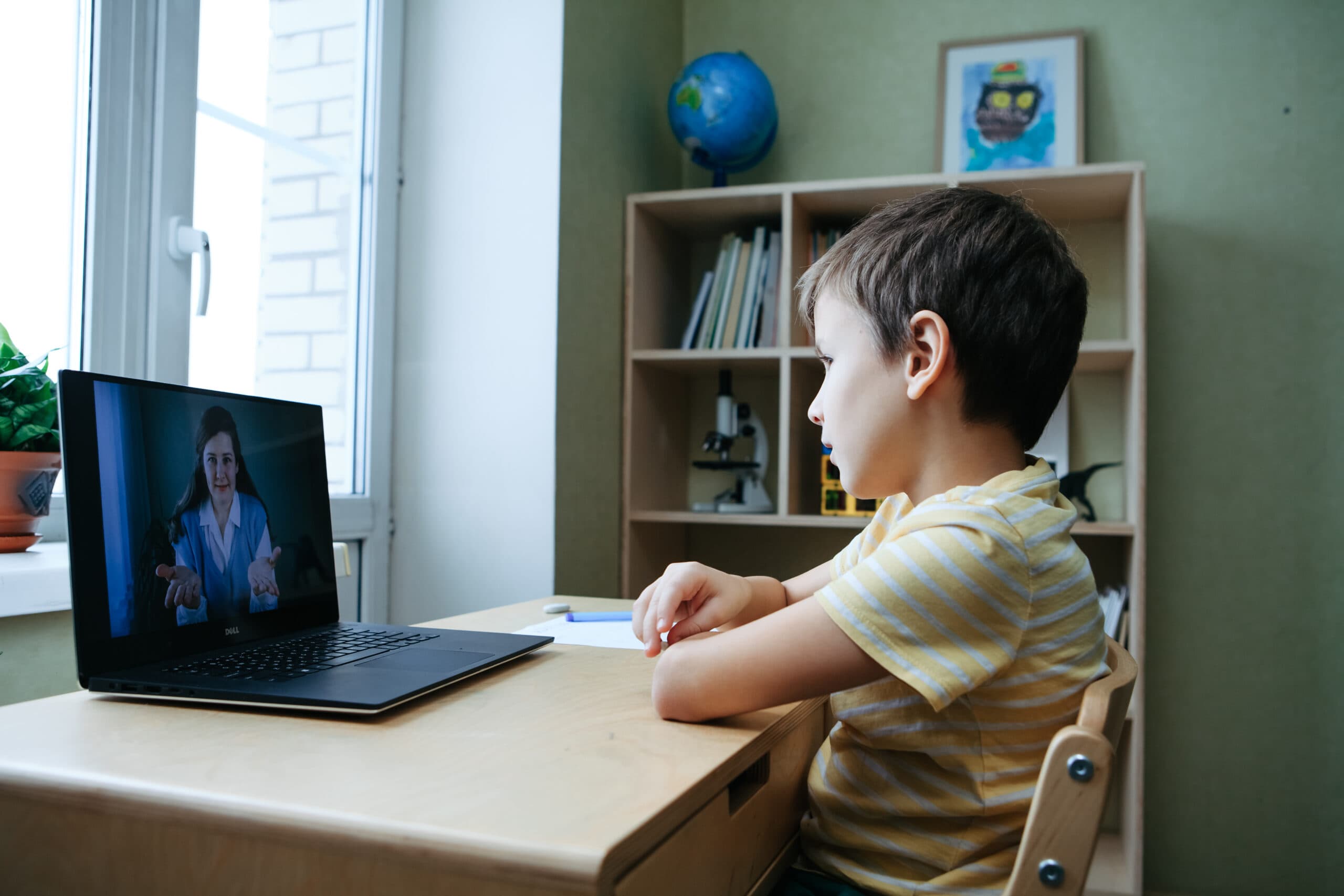 En ung dreng i en gulstribet skjorte sidder ved et skrivebord og ser og lytter til en kvinde i et videoopkald på sin bærbare computer. I baggrunden ses en bogreol og en globus.