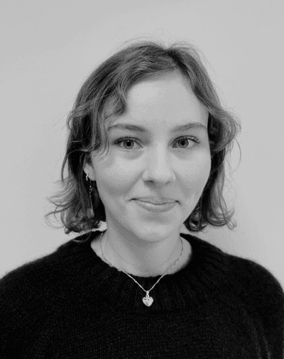 Sort-hvidt portræt af en ung kvinde med kort, bølget hår, iført en mørk trøje og en halskæde med et hjerteformet vedhæng, der poserer foran en ensfarvet baggrund.
