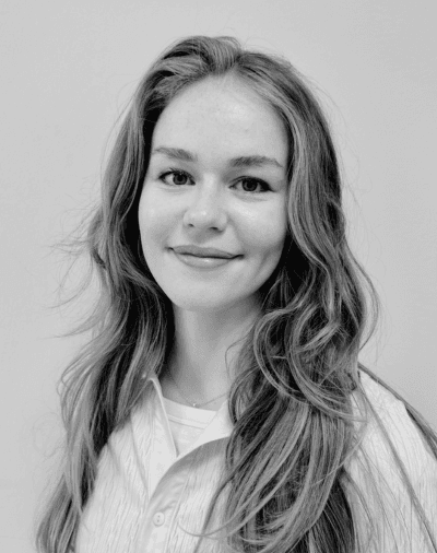 En ung kvinde med langt, bølget hår smiler blidt. Hun har en skjorte med krave på og står foran en enkel, lys baggrund. Billedet er i sort-hvid.