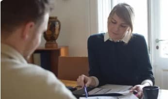 En kvinde sidder ved et bord og gennemgår dokumenter sammen med en mand, der peger på et papir med en kuglepen. De ser ud til at have en fokuseret diskussion i et veloplyst rum.