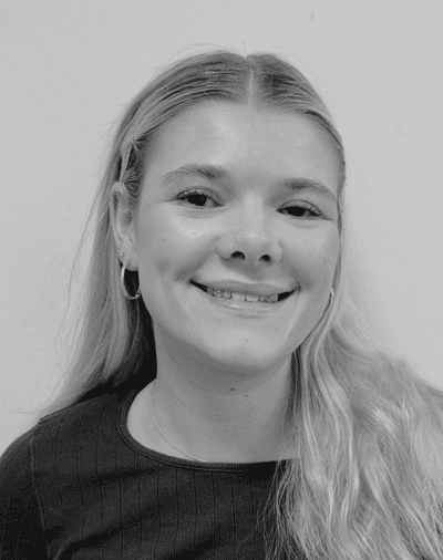 Sort-hvidt foto af en ung kvinde med langt, lyst hår, der smiler. Hun har hoop-øreringe på og en ribbet, mørk top og står foran en ensfarvet lys baggrund.