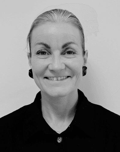 En smilende kvinde med lyst hår, der er sat op, iført mørke øreringe og en mørk skjorte med krave, fotograferet foran en ensfarvet lys baggrund. Sort-hvidt foto.