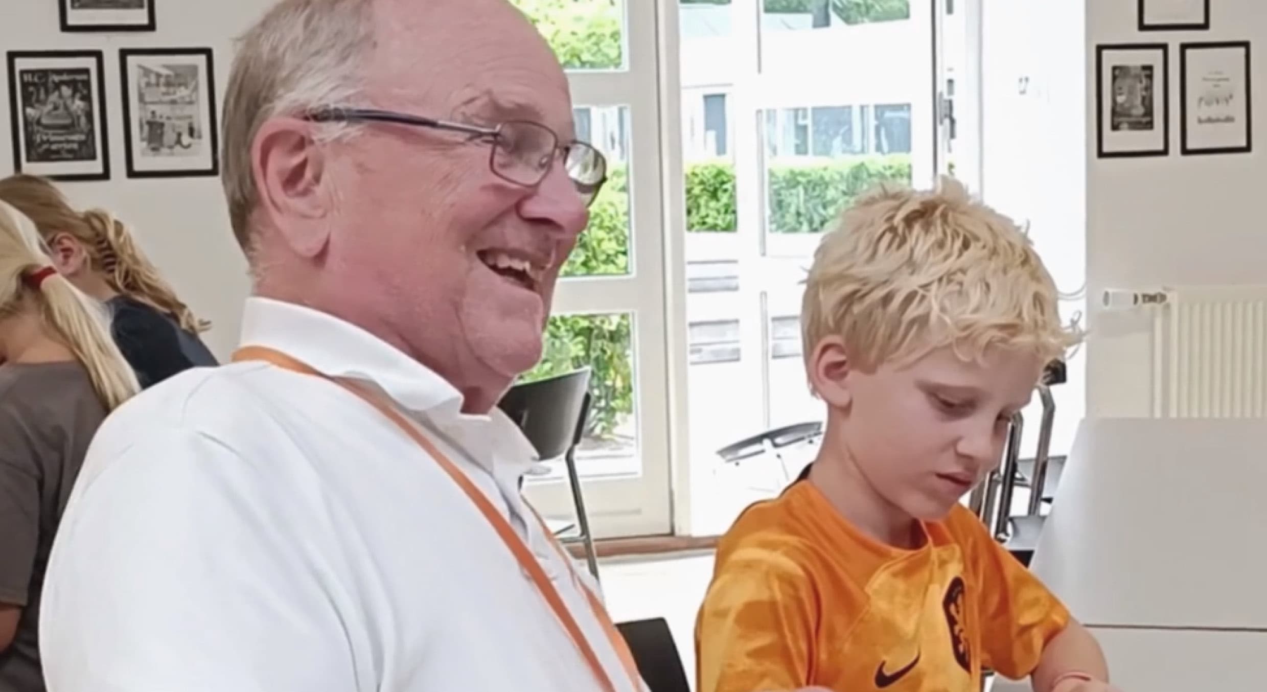 En ældre mand smiler, mens han sidder ved siden af en lille dreng i en orange trøje, der er helt optaget af at tegne ved et bord i et lyst rum.
