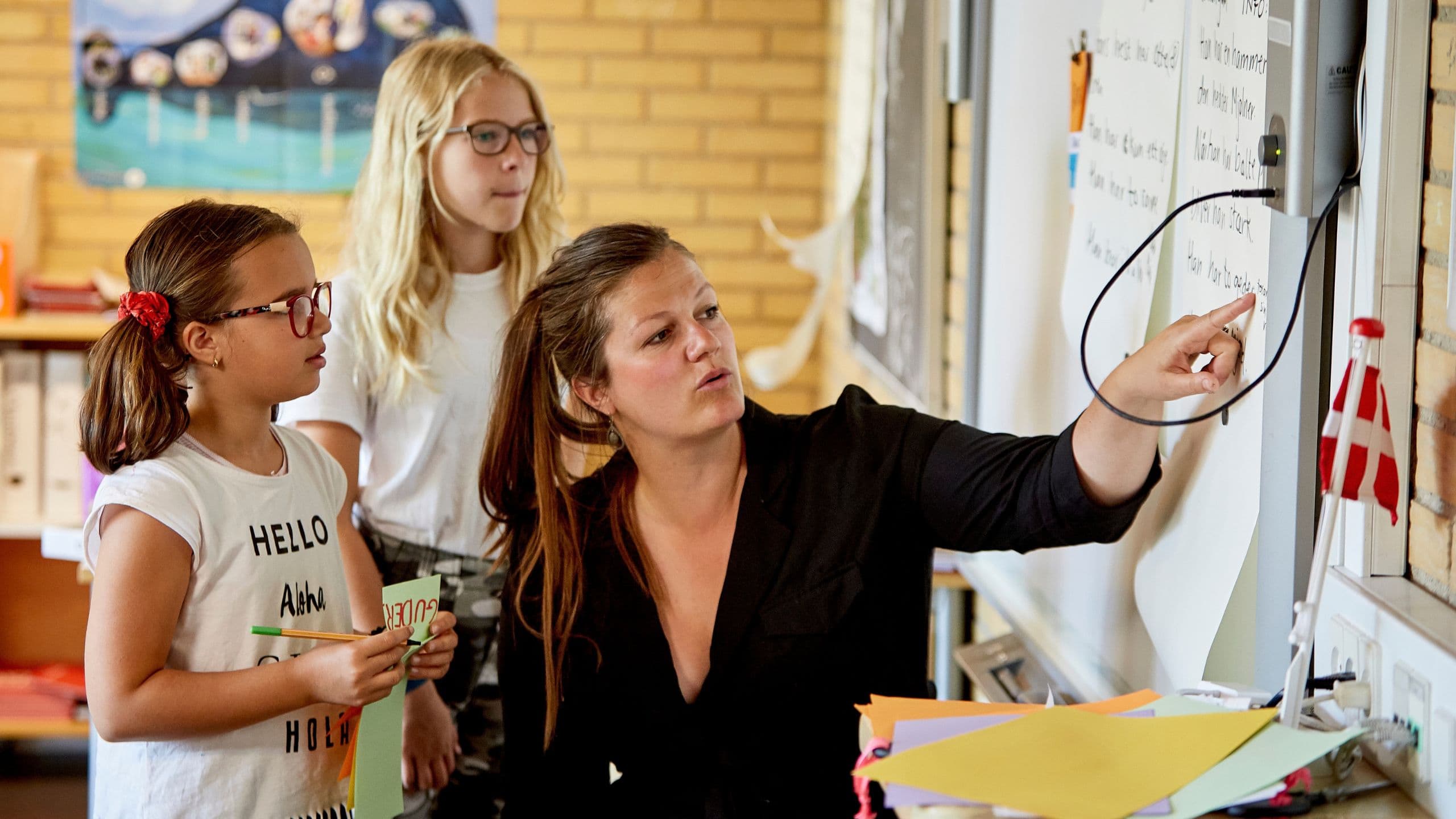 En lærer peger på en tavle, mens to unge elever med briller følger opmærksomt med under en Lær dansk-sommerskole. På skrivebordet i forgrunden ligger der papirer og et lille dansk flag.
