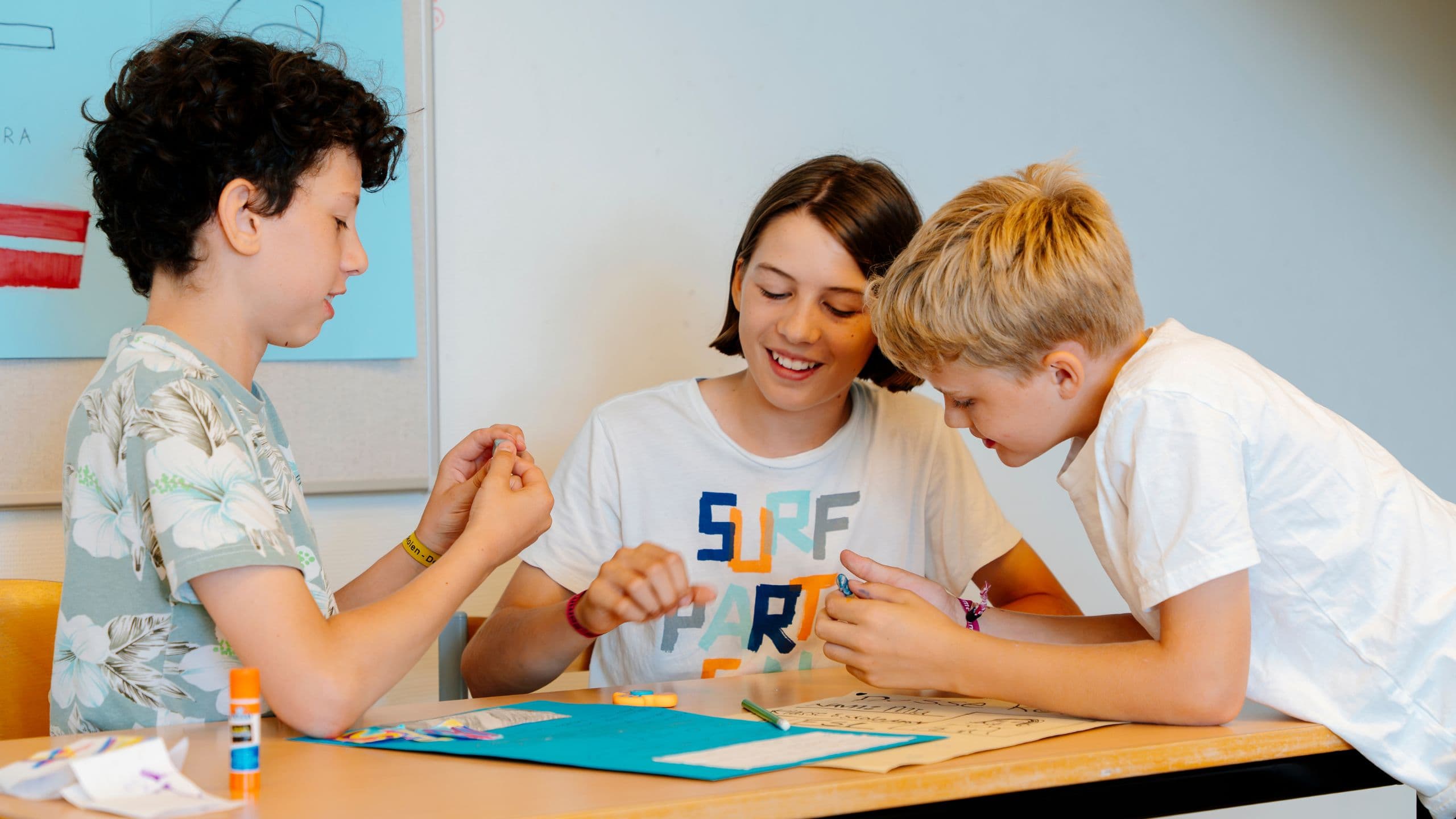 Tre børn sidder ved et bord, smiler og arbejder sammen om et håndværksprojekt med papir og lim på Lær dansk sommerskole Danmark.