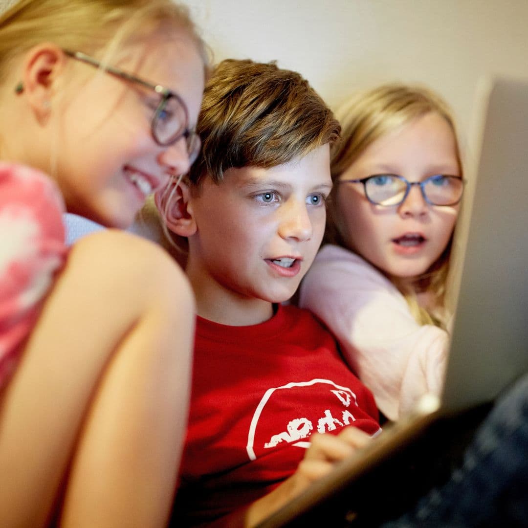 Tre børn sidder tæt sammen og kigger interesseret og begejstret på en bærbar computer – måske er de ved at udforske Lær dansk sommerskole Danmark. De to piger har briller på, og drengen i midten har en rød skjorte på.