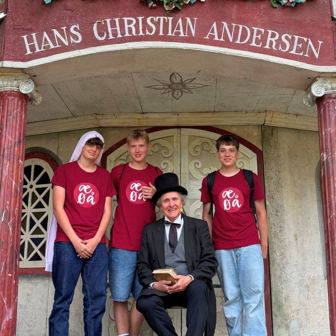 Tre teenagere i ens røde trøjer står ved siden af en mand i sort jakkesæt og høj hat, der holder en bog, og de poserer alle sammen foran Hans Christian Andersen-bygningen på Lær dansk sommerskole Danmark.