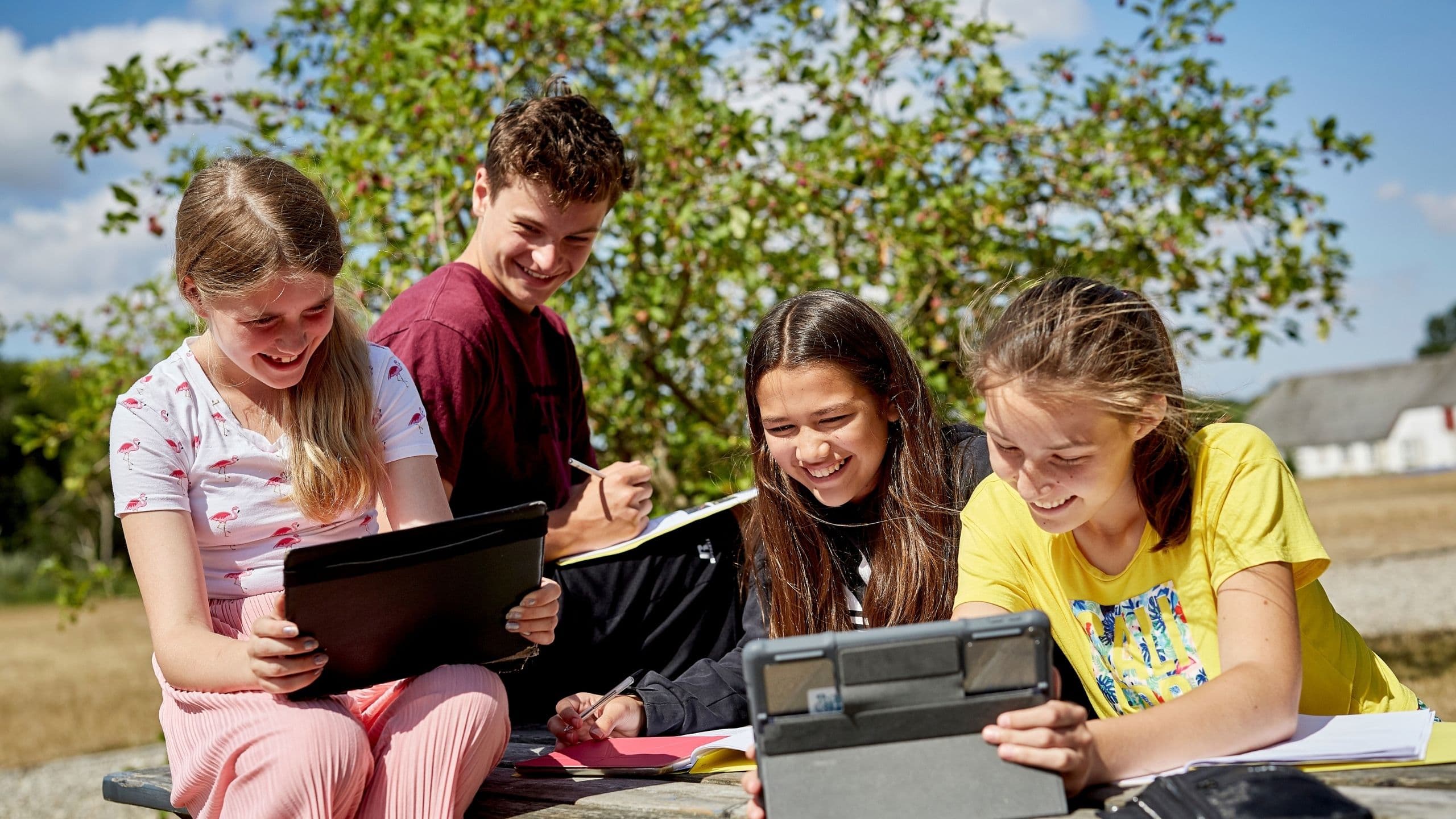 Fire smilende børn sidder udendørs på en bænk, hvor de arbejder sammen med tablets og notesbøger og nyder en solrig dag. I baggrunden ses træer og bygninger på en dansk sommerskole i Danmark.