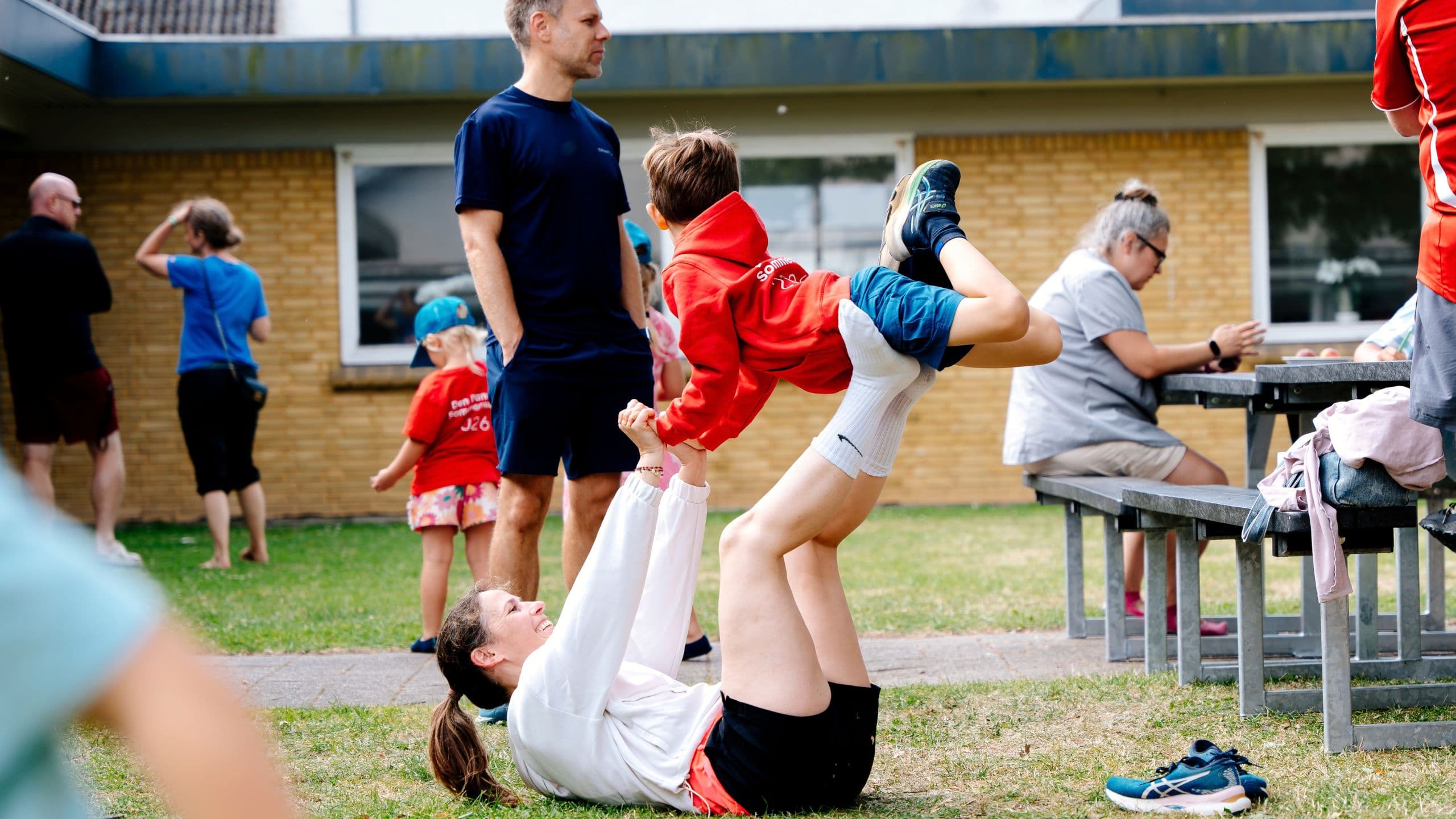 En kvinde på Familiehøjskolen ligger på græsset og løfter et smilende barn i en rød hættetrøje med benene. En voksen står tæt ved, mens andre samles ved en bygning og et picnicbord, klar til at lære dansk sammen.