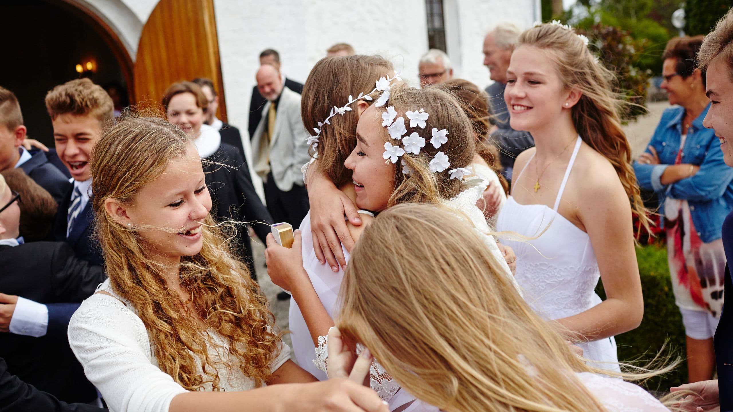 En gruppe unge piger i hvide kjoler omfavner hinanden og smiler uden for en bygning, omgivet af folk, der fejrer konfirmation i Danmark.