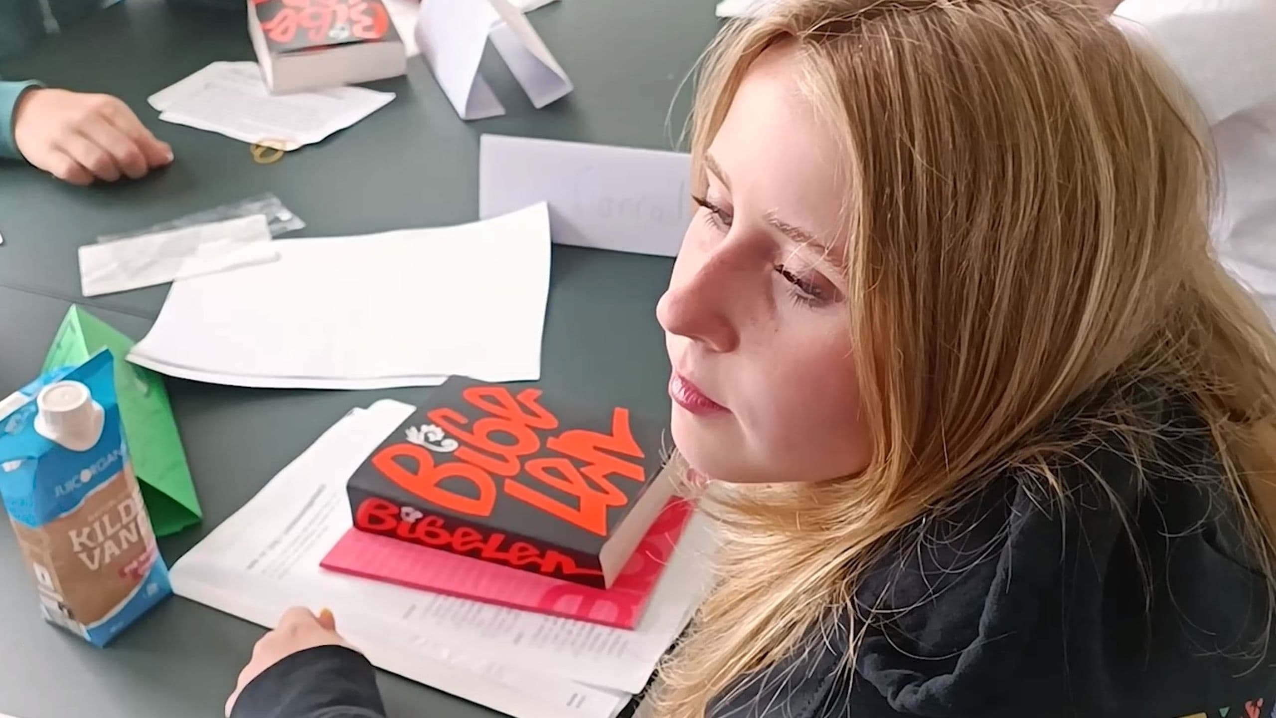En ung kvinde med blondt hår sidder ved et bord med papirer, en blå drikkekarton og to bøger med titlen Bibelen – et billede, der fanger en scene fra en konfirmation i Danmark eller blandt unge udlandsdanskere. Hun er omgivet af andre hænder og bøger.