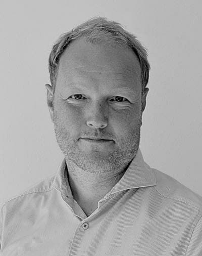 Et sort-hvidt portræt af en mand med kort, lyst hår, et velplejet skæg og et neutralt udtryk, iført en skjorte med krave, foran en ensfarvet baggrund.