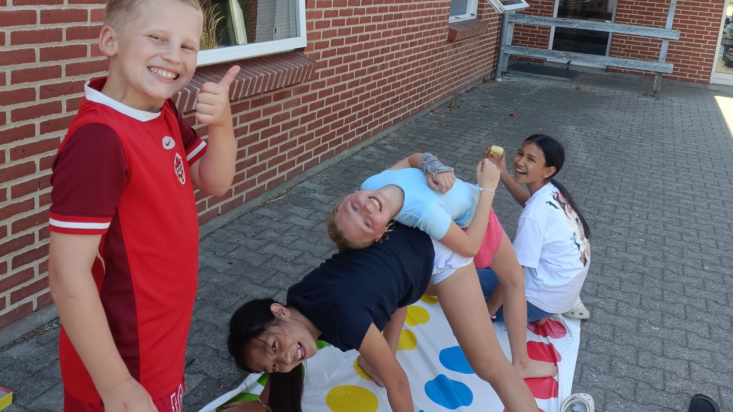 Fire børn leger Twister på en måtte udenfor, hvor de griner og ligger sammenfiltret på Sommerskole for børn i Danmark, mens et andet barn i en rød trøje står tæt ved og smiler og giver tommelfingeren op ved siden af en murstensbygning.