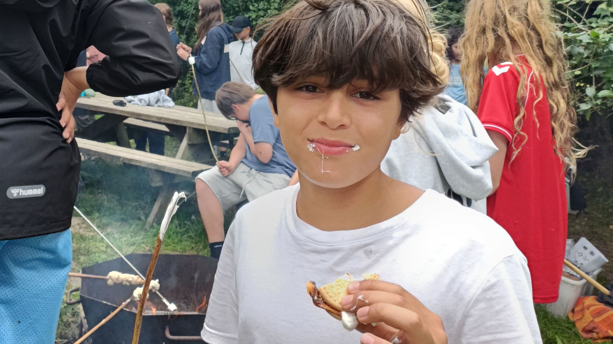 En dreng med brunt hår spiser en s’more med en skumfidus på læberne, mens han står ved et lejrbål på Sommerskole for børn i Danmark. Andre børn rister skumfiduser og hygger sig i den afslappede udendørs atmosfære.