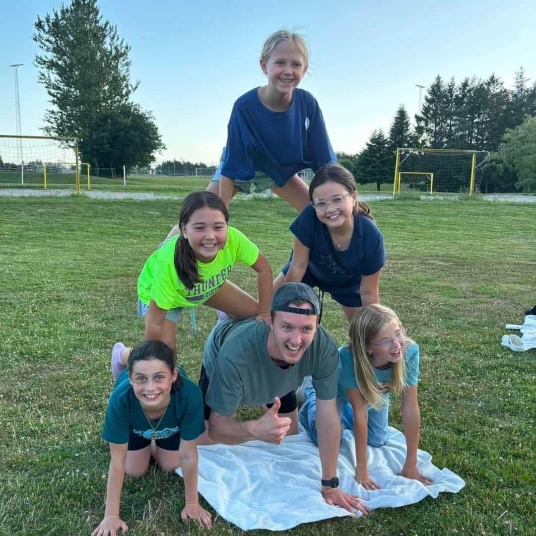 Seks personer – fem børn og en voksen – danner en menneskepyramide på græsset ved Sommerskole i Danmark, med fodboldmål og træer i baggrunden. Alle smiler, og der ligger et håndklæde udbredt under dem.