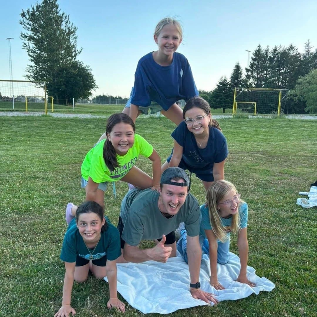 Seks personer – fem børn og en voksen – danner en menneskepyramide på græsset ved Sommerskole i Danmark, med fodboldmål og træer i baggrunden. Alle smiler, og der ligger et håndklæde udbredt under dem.