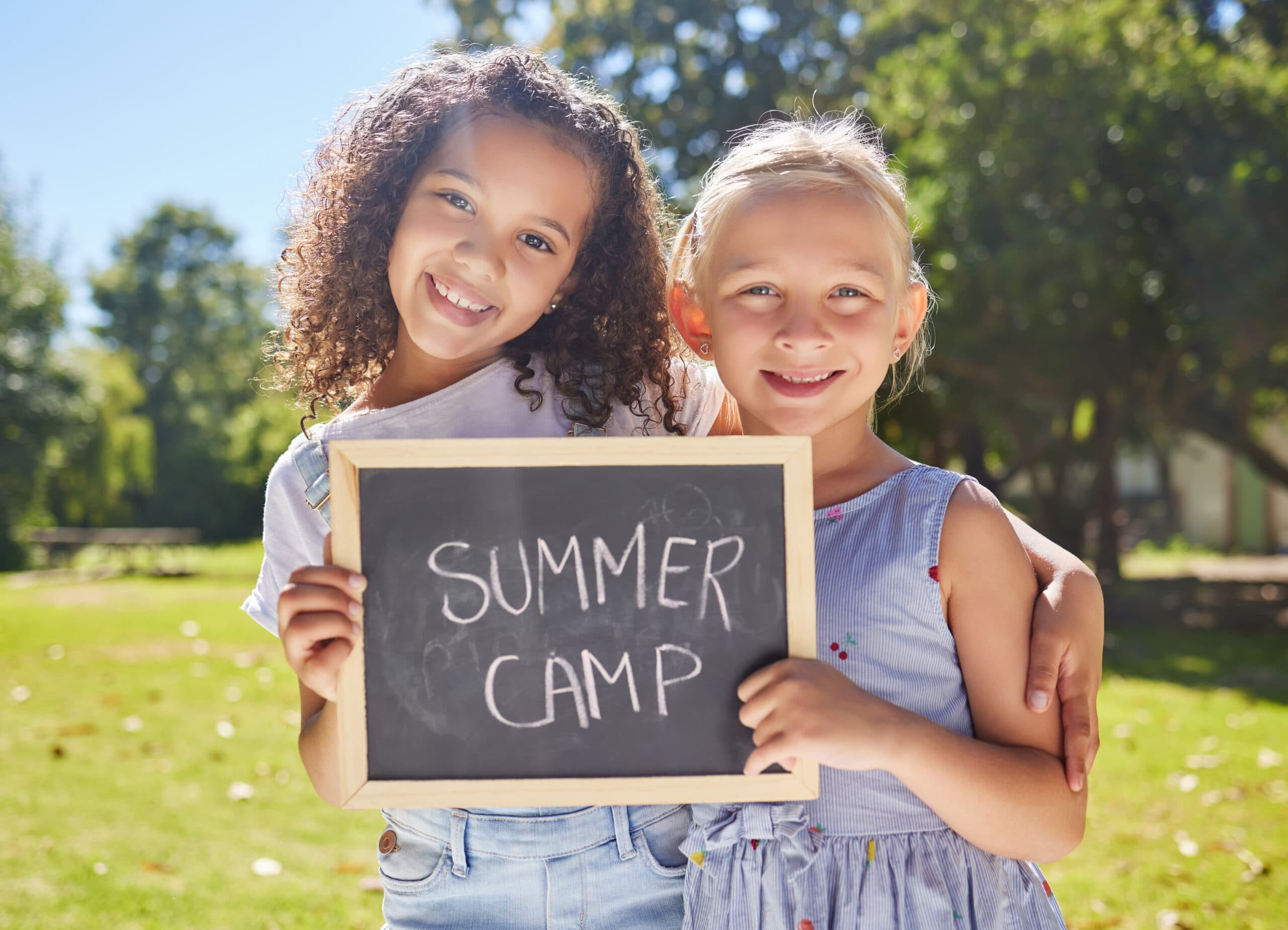 To smilende piger står udendørs på en solskinsdag med en arm om hinanden og holder et kridttavleskilt med teksten SUMMER CAMP. Træer og græs er synlige i baggrunden.