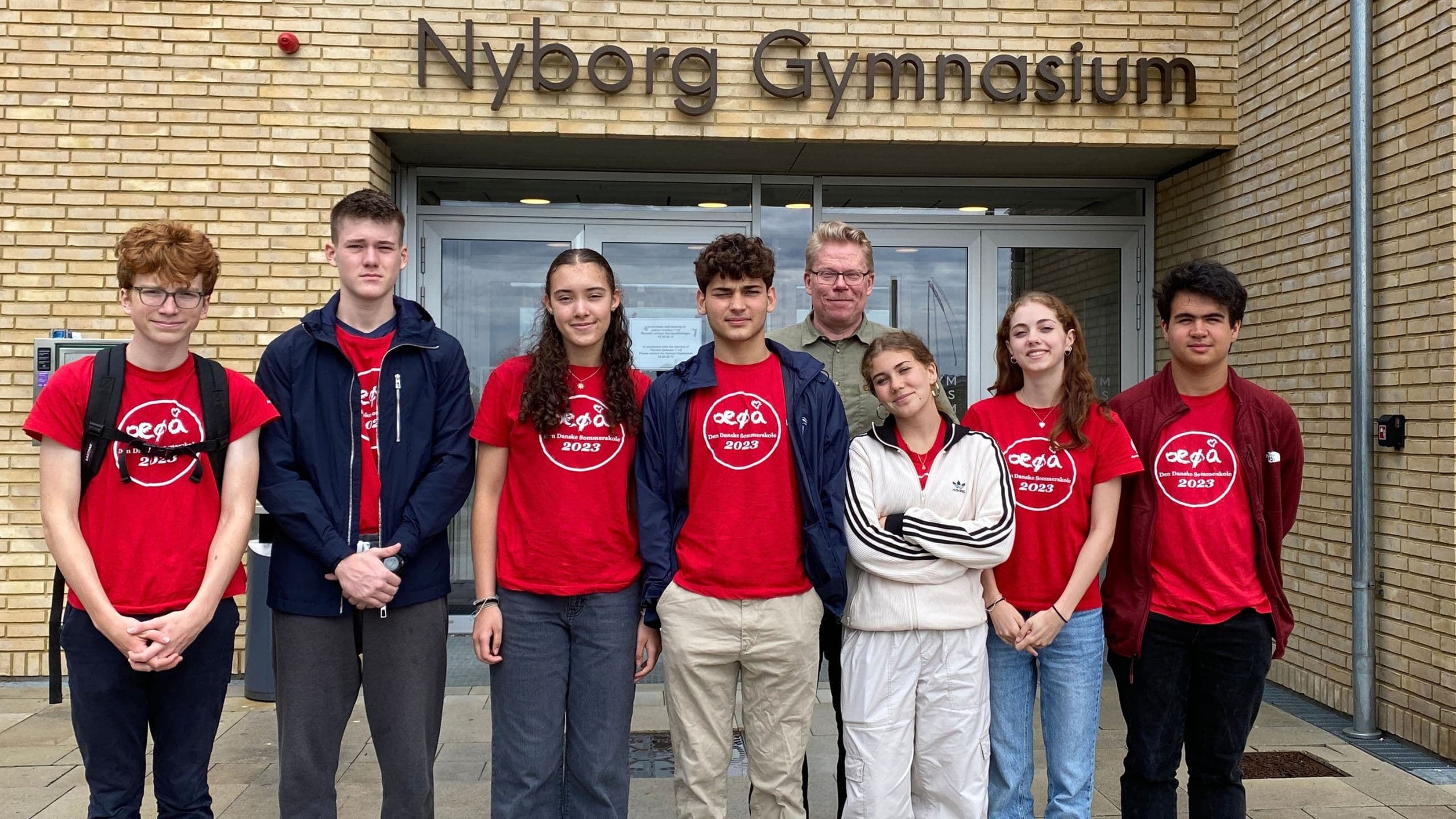 Otte personer står foran Nyborg Gymnasium med dets gule murstensfacade – syv i ens røde T-shirts og én i et beige træningssæt – klar til at deltage i Flyt til Danmark ung og begynde deres fælles rejse.