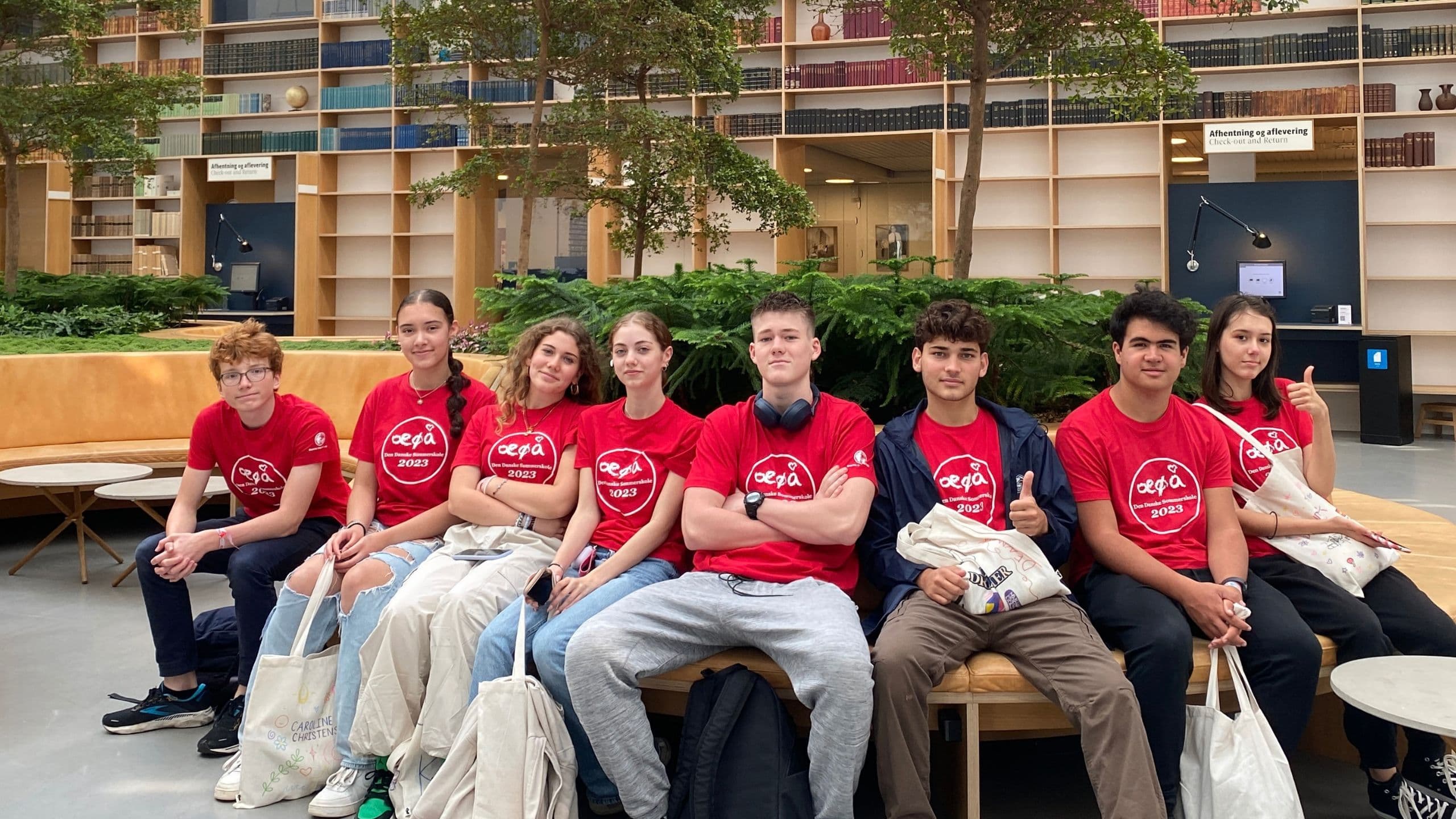 Otte teenagere iført ens røde T-shirts sidder på en buet bænk i et moderne bibliotek med stofposer i hænderne. Bag dem danner frodige grønne planter og bogreoler rammen om denne flytning til Danmark – en ung oplevelse.