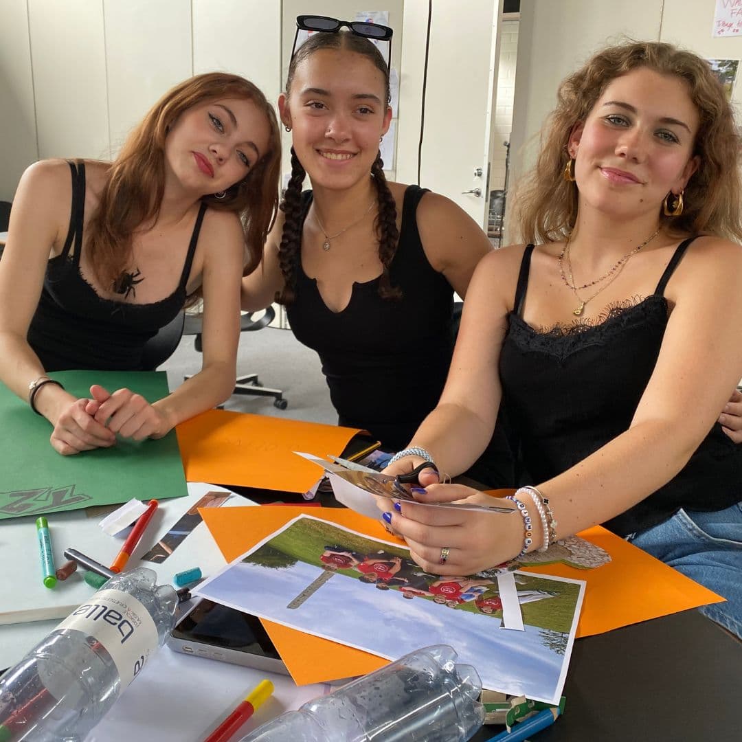 Tre unge kvinder sidder ved et bord med papirer, fotos, saks og kuglepenne – og smiler, mens de arbejder på et skoleprojekt. Der ligger vandflasker og hobbyartikler spredt rundt omkring; måske planlægger de at flyve til Danmark som unge.
