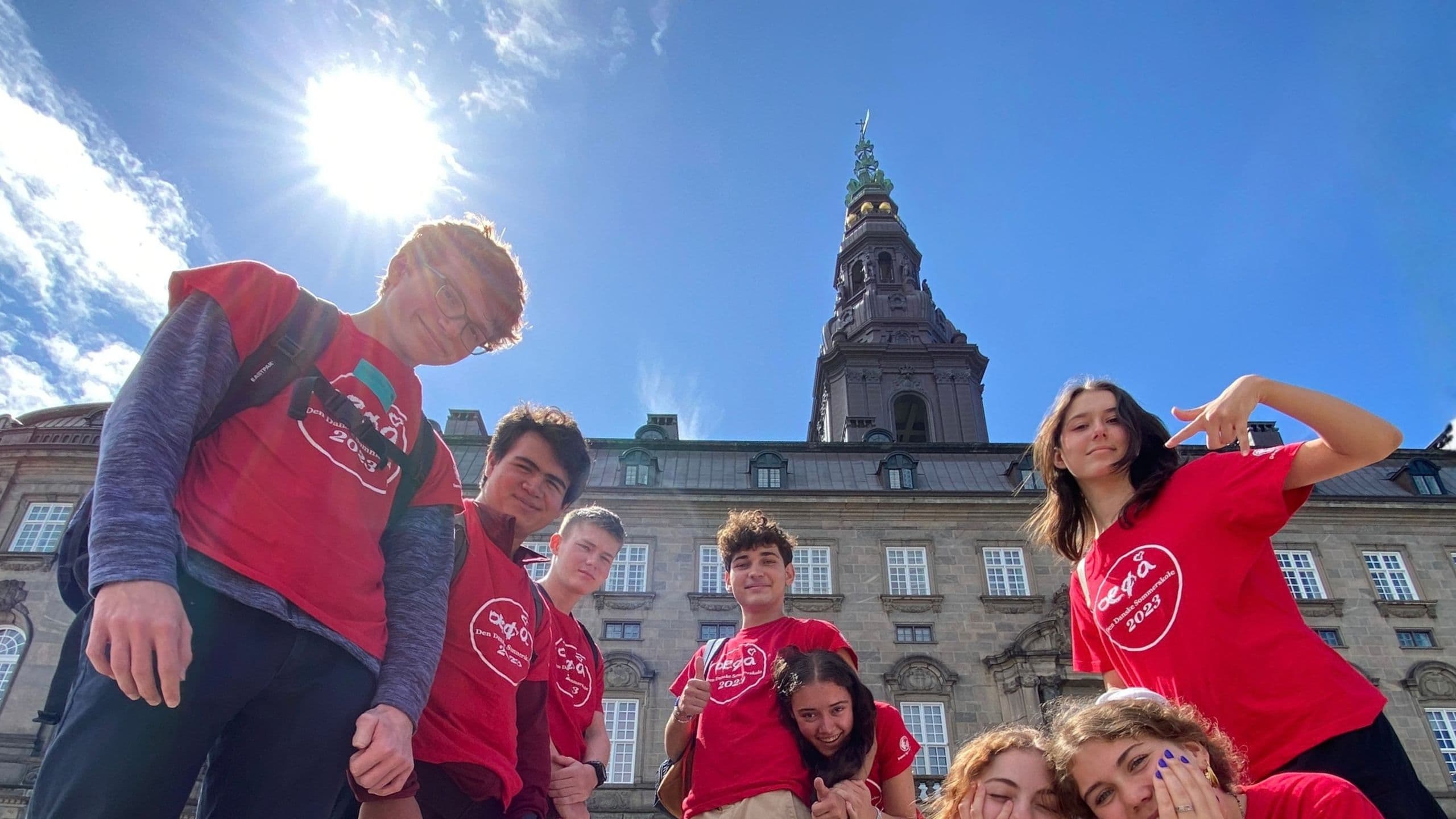 En gruppe unge i røde trøjer poserer og smiler foran en historisk bygning med et højt tårn under en klar, solrig himmel.