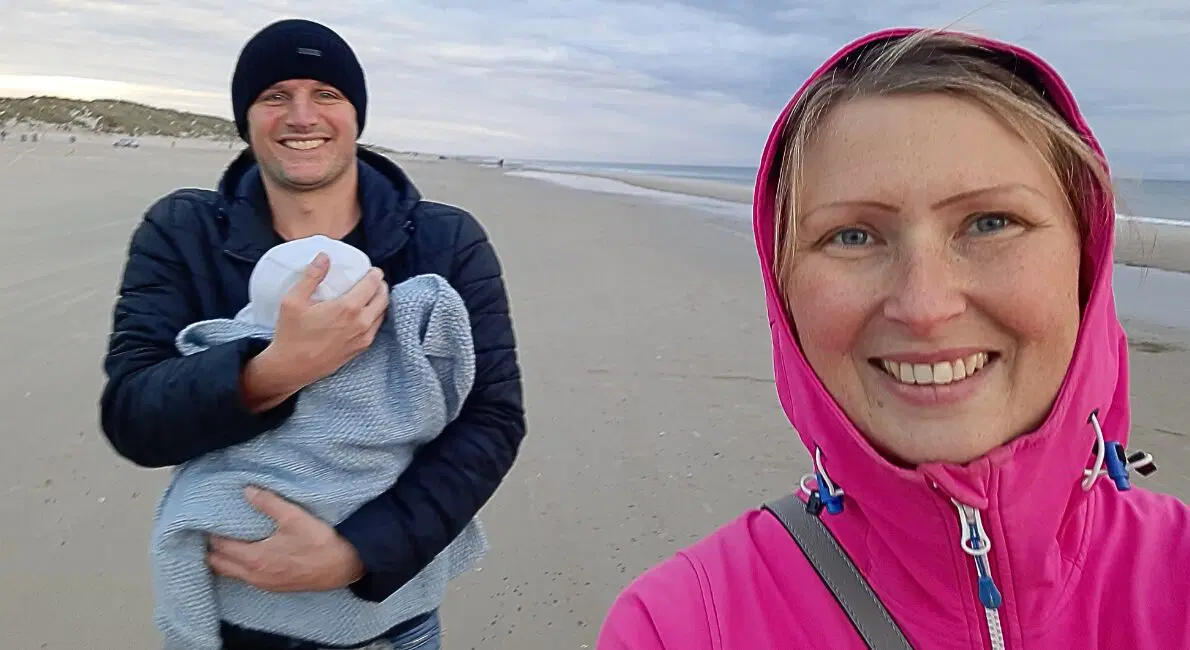 En kvinde i en pink jakke tager en selfie på en strand og smiler til kameraet. Bag hende holder en mand i mørk jakke og hue en baby indsvøbt i et tæppe. Stranden og den overskyede himmel ses i baggrunden.