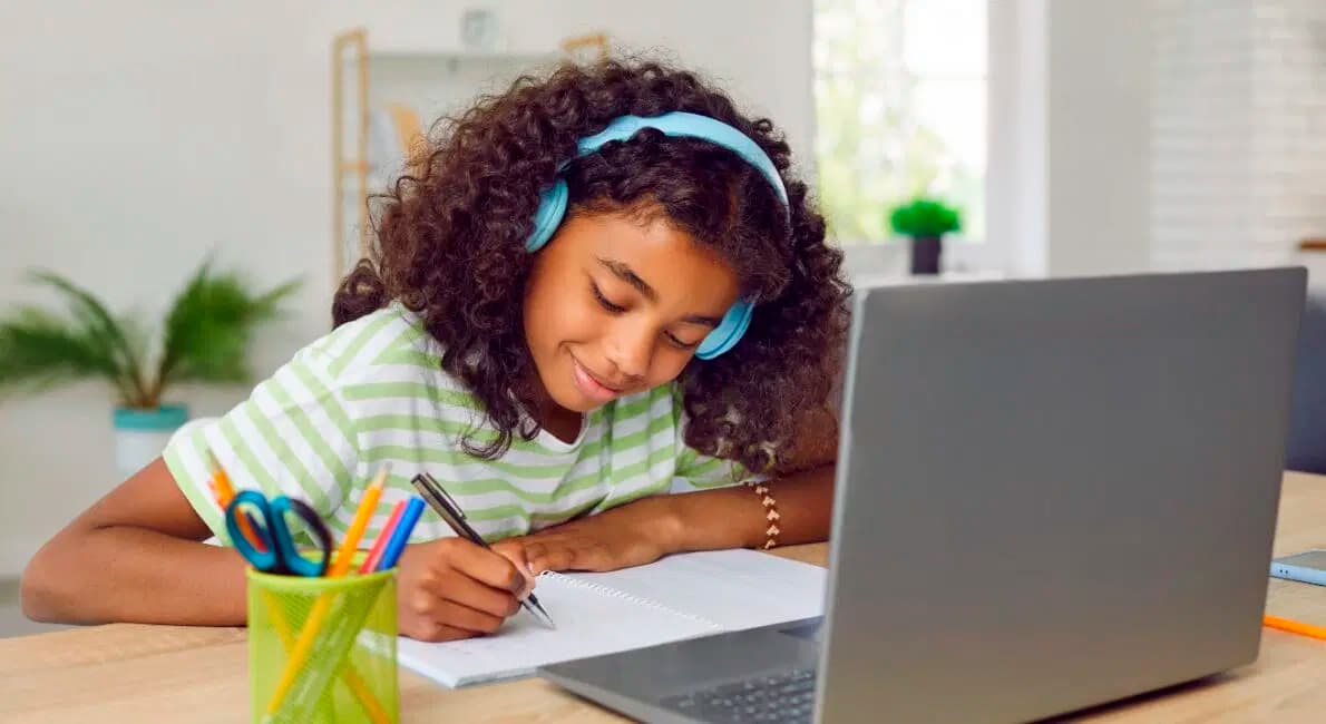 En ung pige med krøllet hår bærer blå hovedtelefoner og en stribet skjorte og smiler, mens hun skriver i en notesbog ved siden af en bærbar computer på et skrivebord med farverige kuglepenne og en plante i baggrunden.