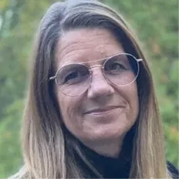 En kvinde med glat, lysebrunt hår og runde briller smiler blidt. Hun er udendørs med en sløret grøn baggrund.