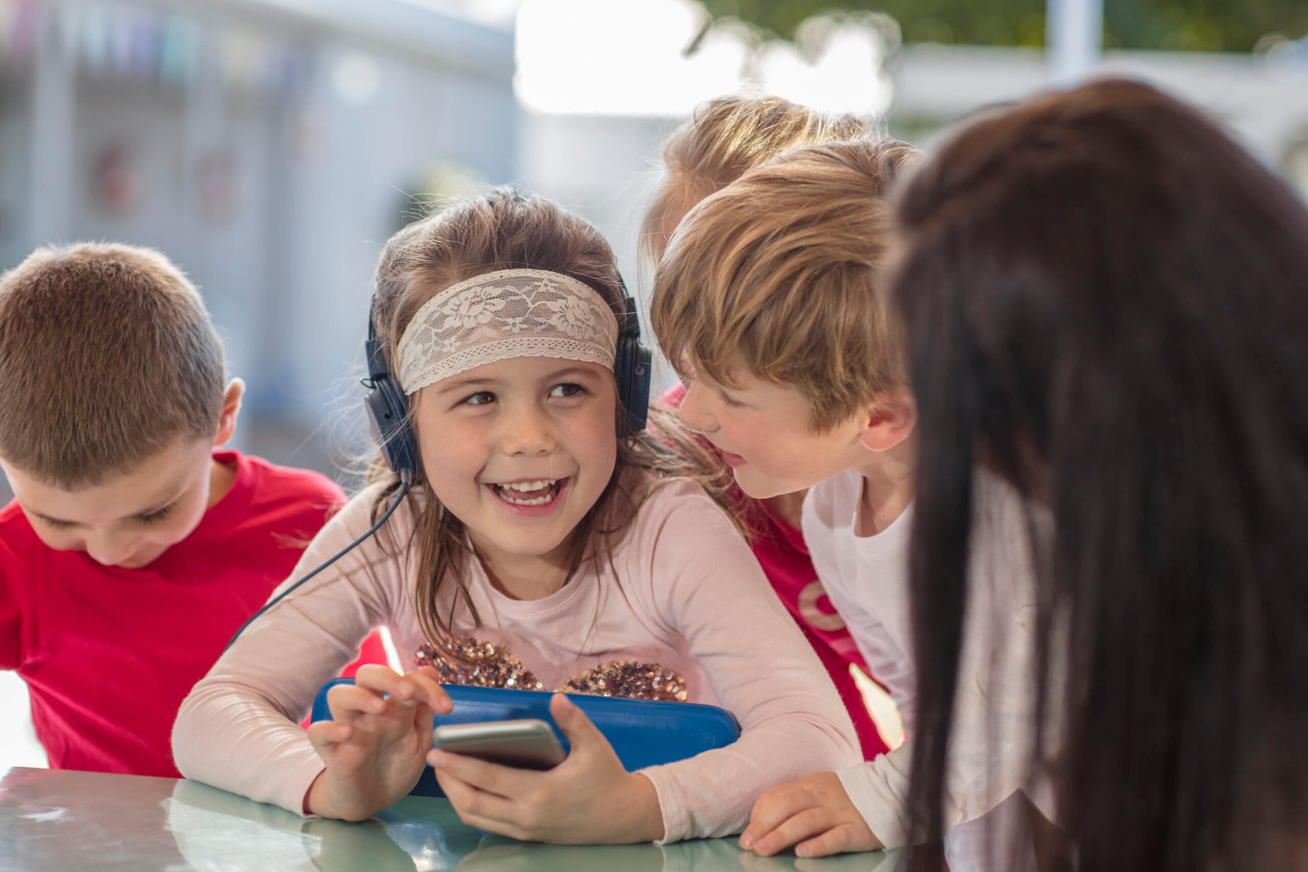En gruppe små børn samles omkring et bord udenfor. En pige i midten har hovedtelefoner på, smiler og holder en tablet, mens de andre læner sig tæt op ad, engagerede og muntre.