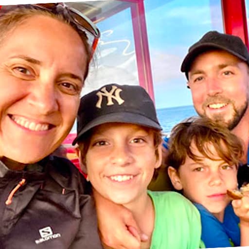 En kvinde og en mand smiler til en selfie med to små drenge foran et vindue med havudsigt. Den ene dreng har en sort Yankees-kasket på; gruppen virker glad og tæt sammen.