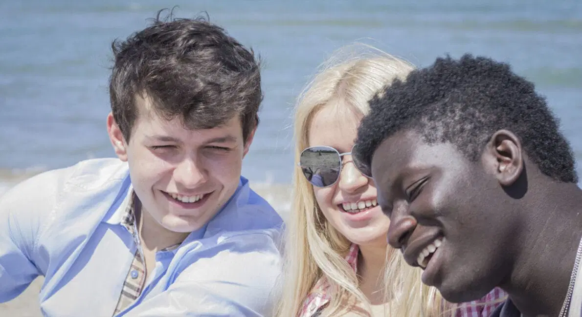Tre venner, to mænd og en kvinde, smiler og poserer til en selfie på stranden med havet i baggrunden. Kvinden har solbriller på. Vejret ser ud til at være solrigt og lyst.